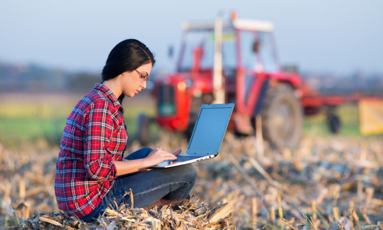 Young woman with laptop sitting on the bale on the field. Tractor in background